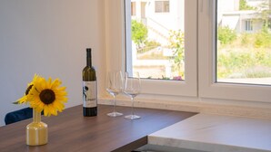 Uma mesa com uma garrafa de vinho e duas taças, um vaso com um girassol, e vista para casas através de uma janela.
