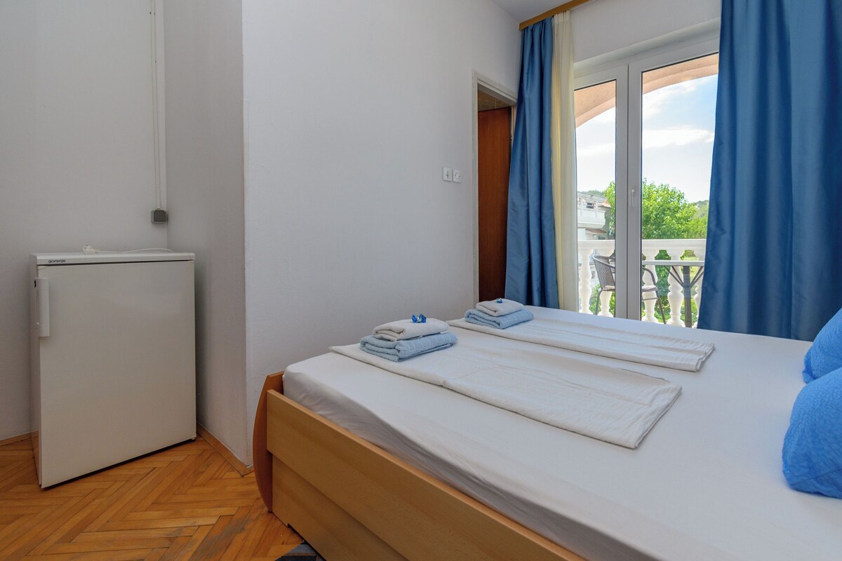 A bedroom with a bed, a small white refrigerator, and a balcony with blue curtains.