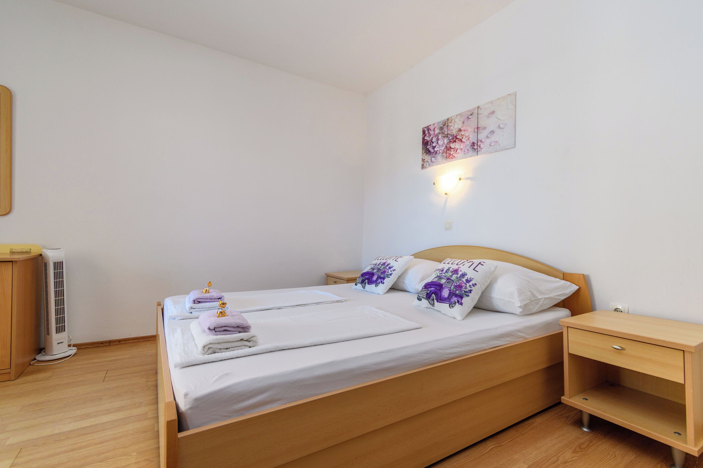 A neatly made bed with white linens and decorative pillows, a bedside table, a wall-mounted light, and a radiator.