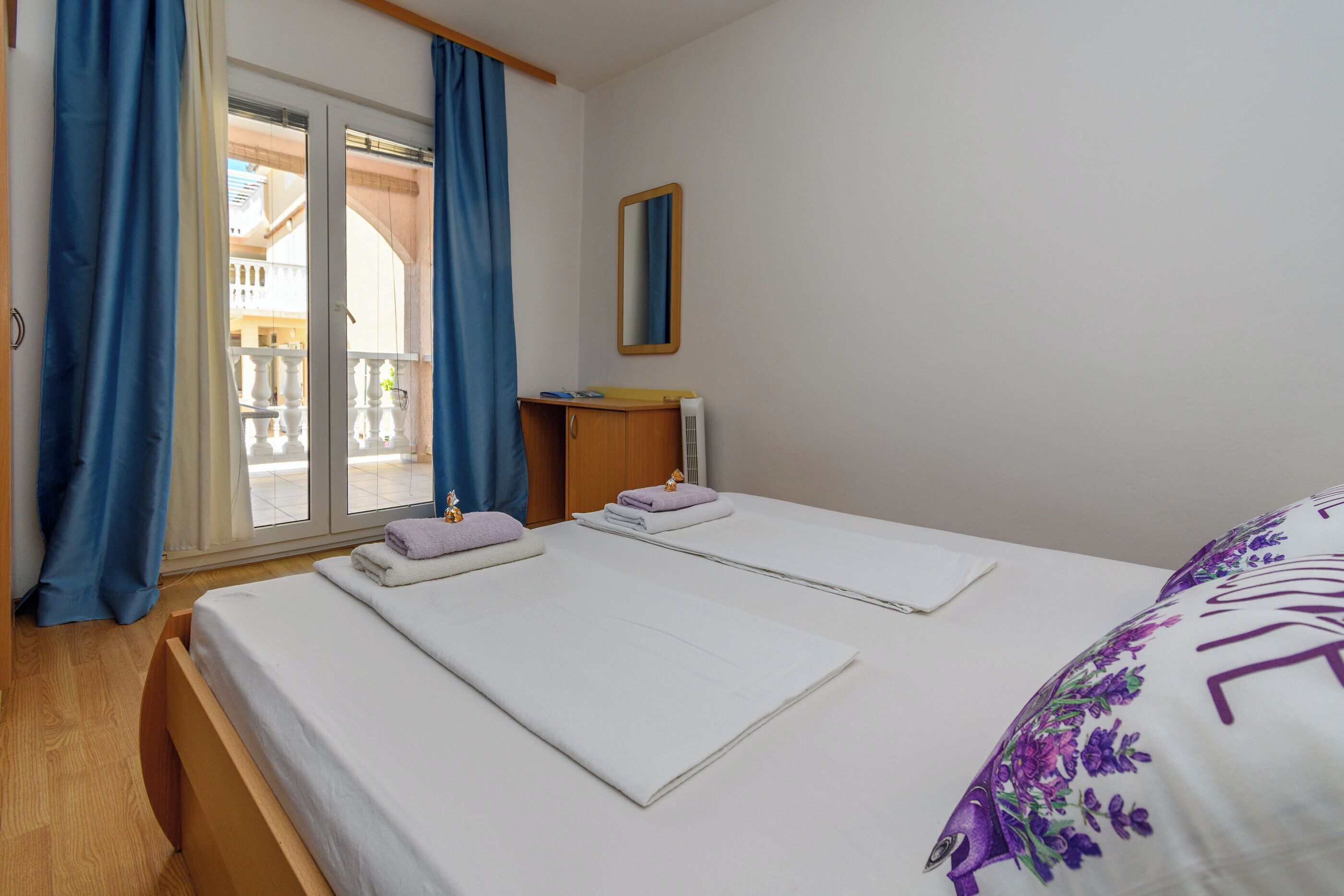 A hotel room with two beds, a wooden wardrobe, a small table, a mirror, and a window with blue curtains.