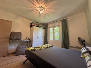 Ein Schlafzimmer mit einem Bett, einem Kleiderschrank, einem Schreibtisch mit Stuhl, einem Fenster mit Vorhängen und einer Deckenleuchte.