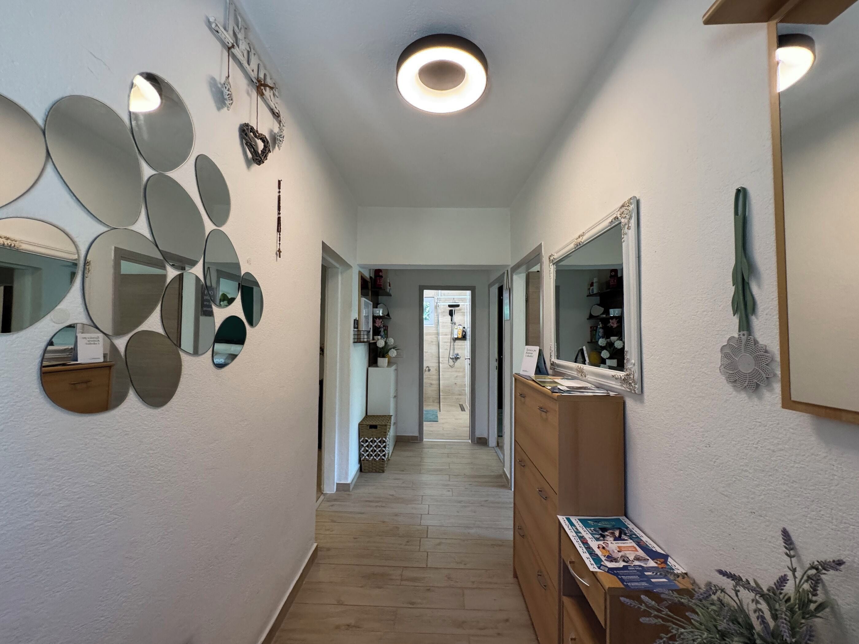 A hallway with a wooden floor, a wooden dresser with a magazine, and a mirror with a circular light.