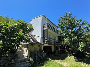Ein zweistöckiges Gebäude mit Balkon, umgeben von Grünpflanzen und einer Steinmauer.