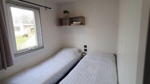 Acomodação pequena, um quarto individual com duas camas, uma janela e uma prateleira com uma planta.