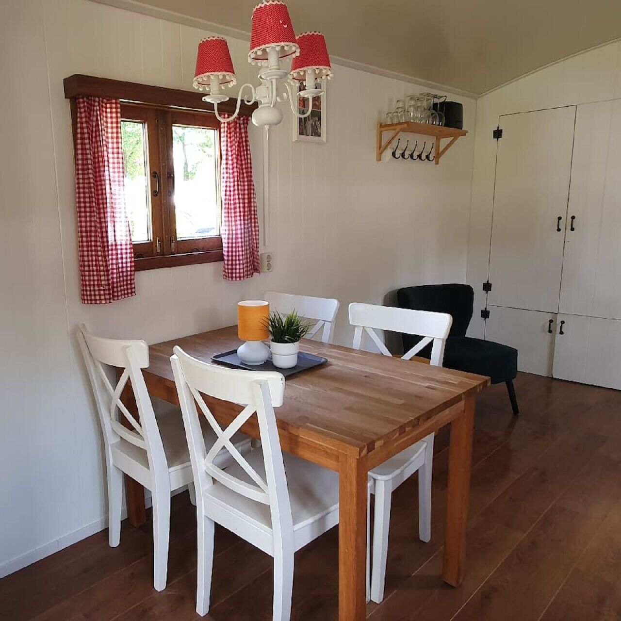 Ein Essbereich mit einem Holztisch, weißen Stühlen, einem rot-weißen karierten Fenster und einer Kristallleuchtre.'