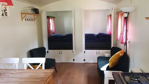 A compact kitchen with a dining area, a stove, and two beds with curtains. - Verblijf in een unieke Pipowagen | De Otterberg (Wijster)