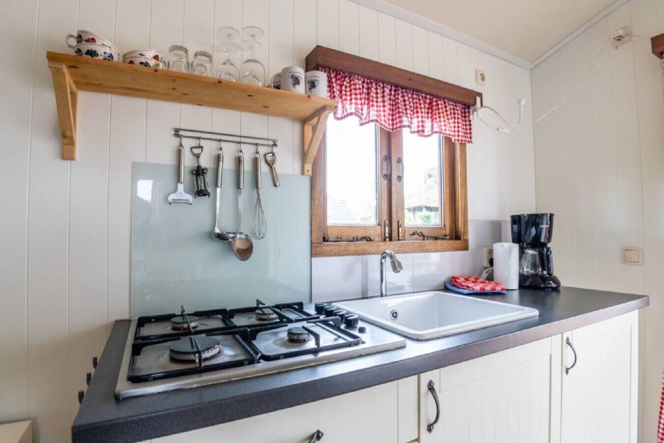 Een keuken met een gasfornuis, een spoelbak en een raam met een rood-wit geruit gordijn.