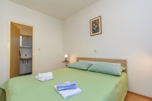 A room with a bed, a nightstand with a lamp, a wall-mounted picture, and a door leading to a bathroom. - Apartments Mande (Rab)