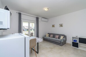 Ein Wohnzimmer mit einer Couch, einem Tisch und einem Sessel. Es gibt ein Fenster mit Vorhängen, eine Klimaanlage und ein Wandregal.