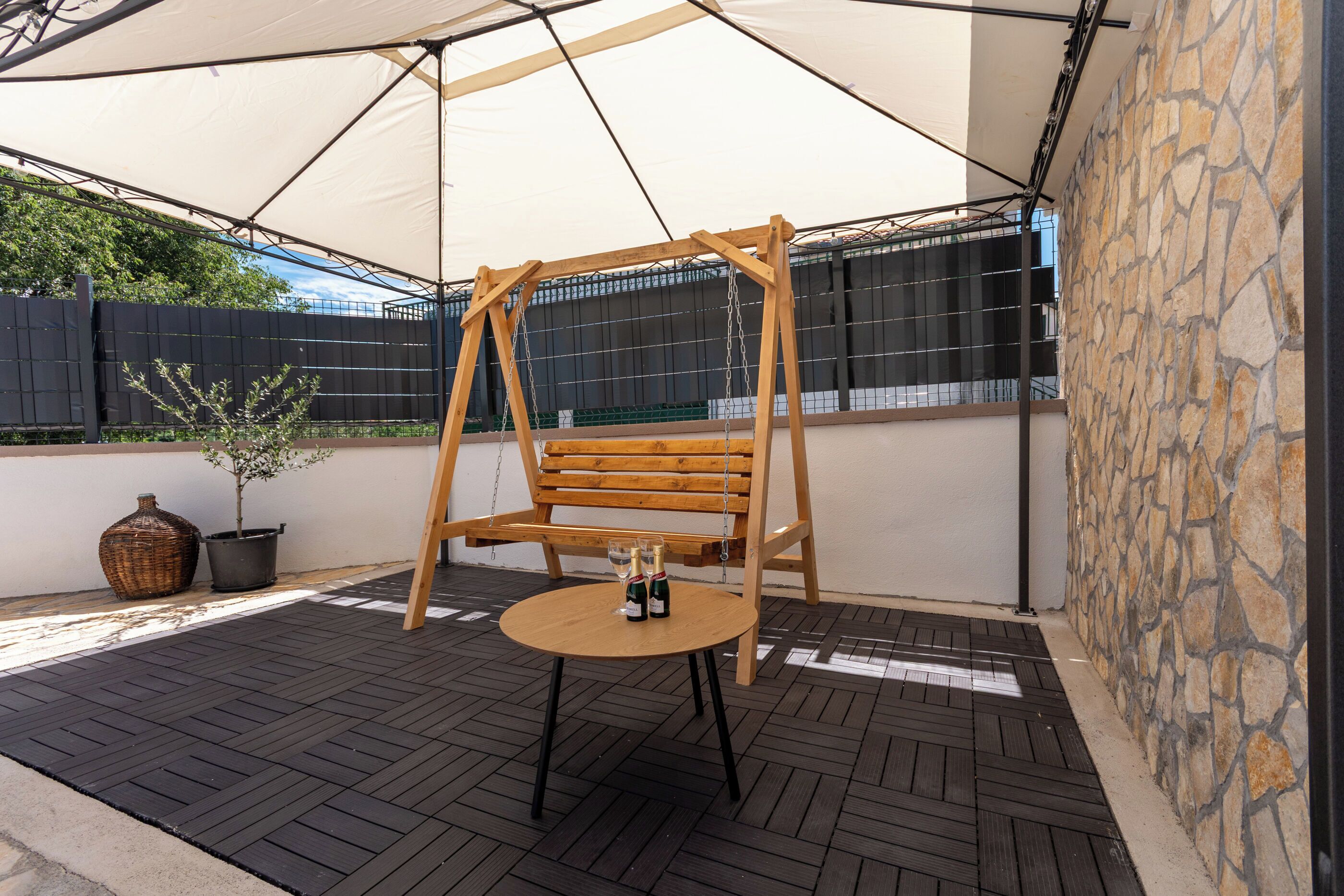 A patio with a swing, a round table with two bottles, a potted plant, and a stone wall.