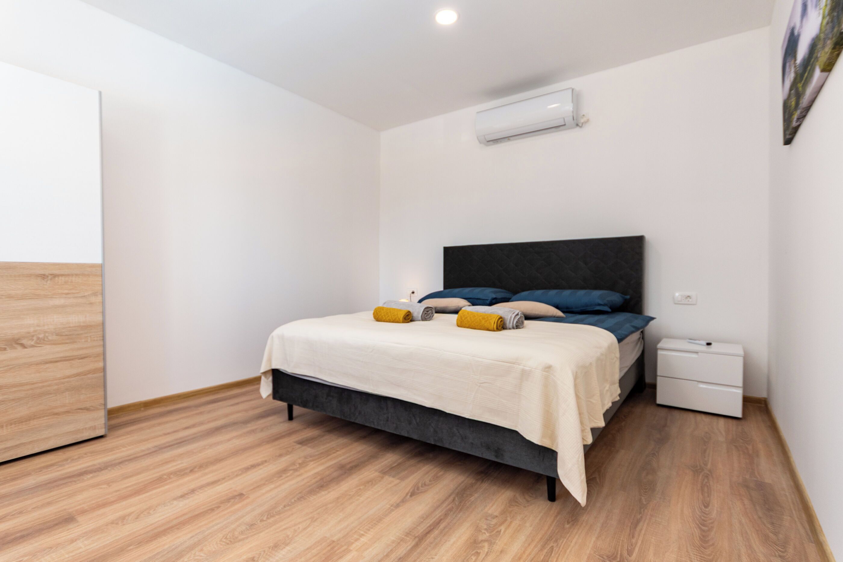 A bedroom with a bed, bedside tables, an air conditioner, and a wooden floor.