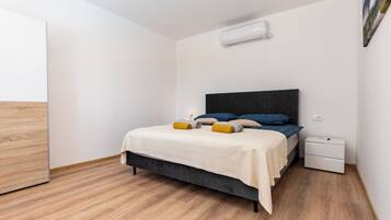 A bedroom with a bed, bedside tables, an air conditioner, and a wooden floor.
