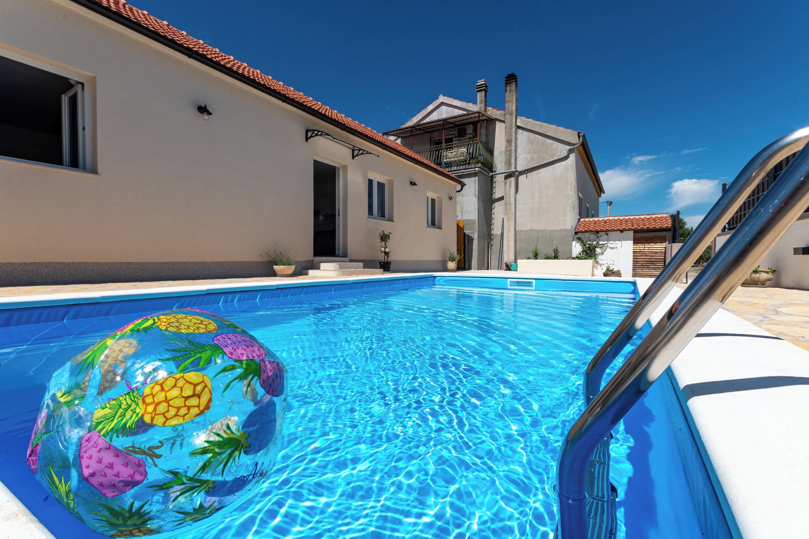 A swimming pool with a pineapple inflatable float, a metal pool ladder, and a building with a tiled roof in the background.