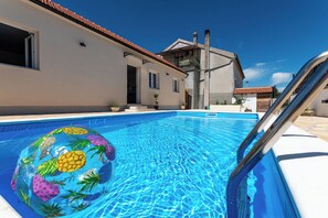 A swimming pool with a pineapple inflatable float, a metal pool ladder, and a building with a tiled roof in the background. - House Mirakul Near Krka With Private Pool (Skradin)