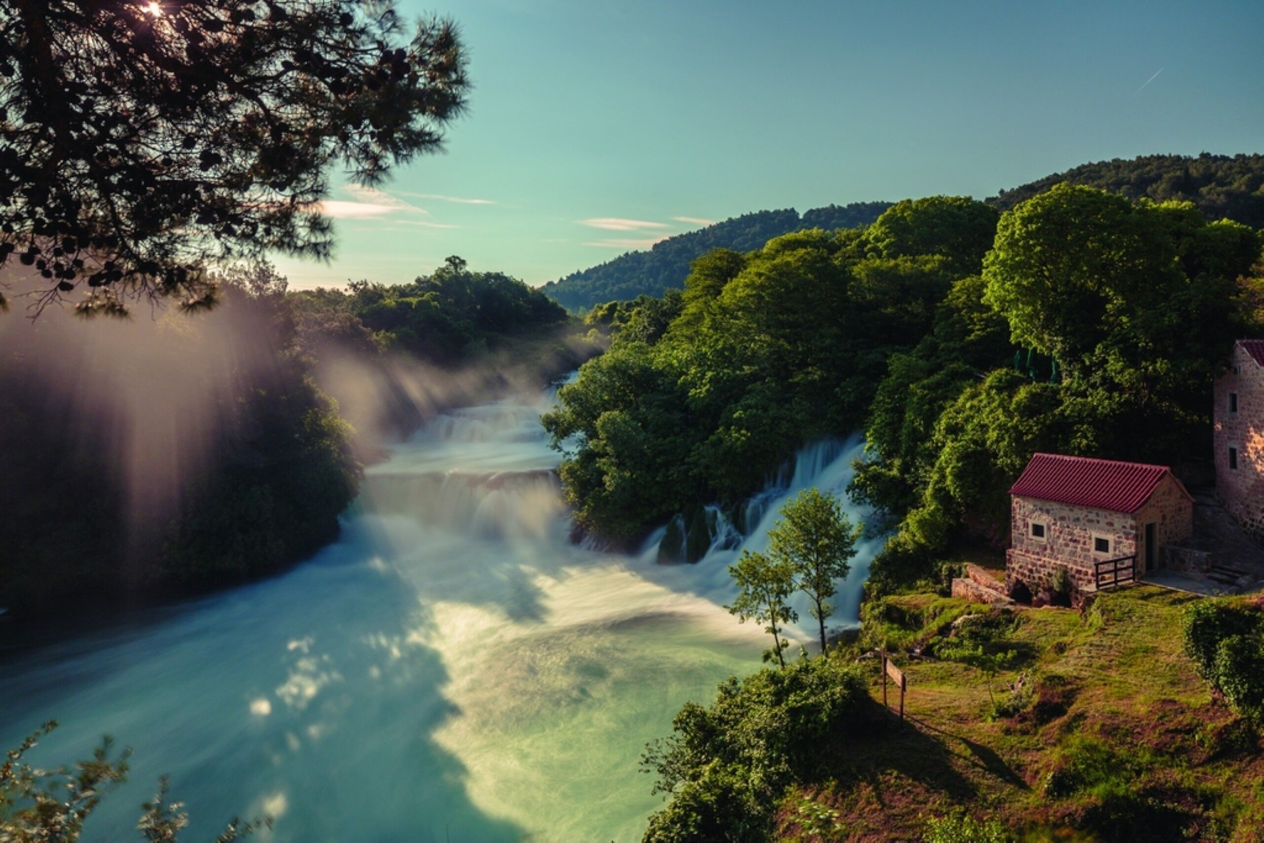 Uma cascata, vegetação exuberante e um edifício de pedra com telhado vermelho.