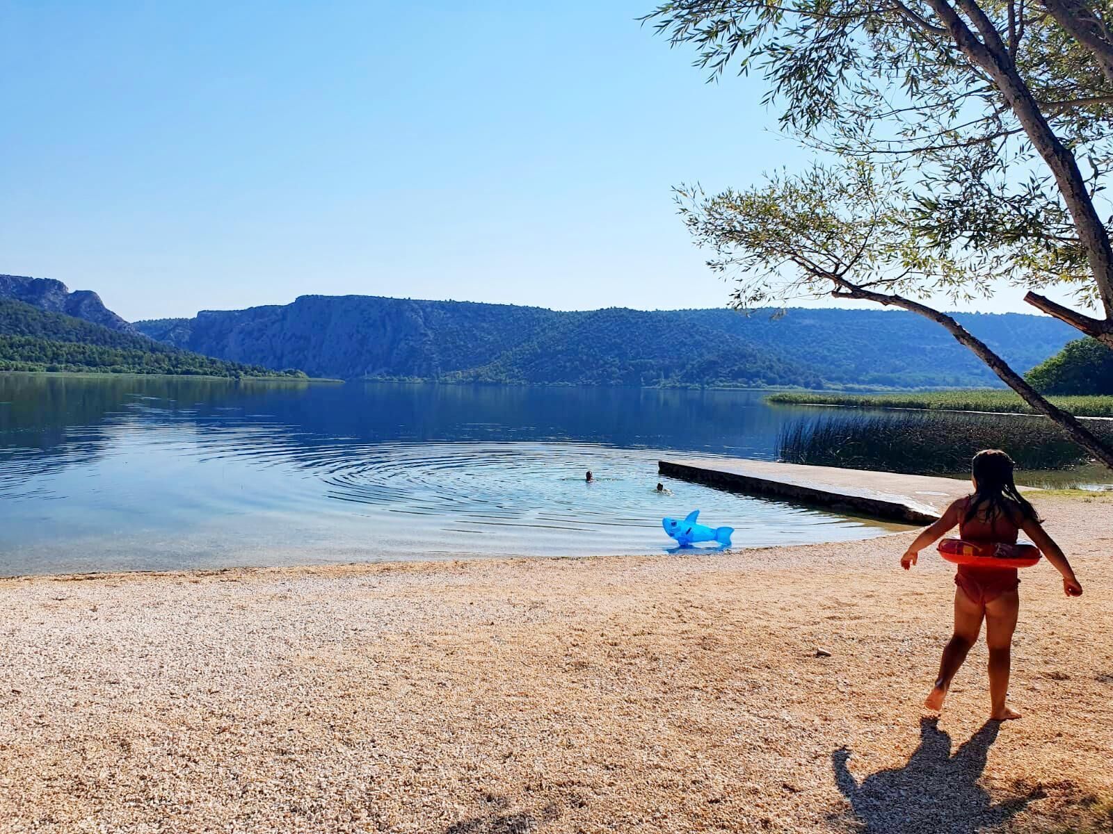 A person standing by a lake with a pebbled shore and a dock extending into the water.