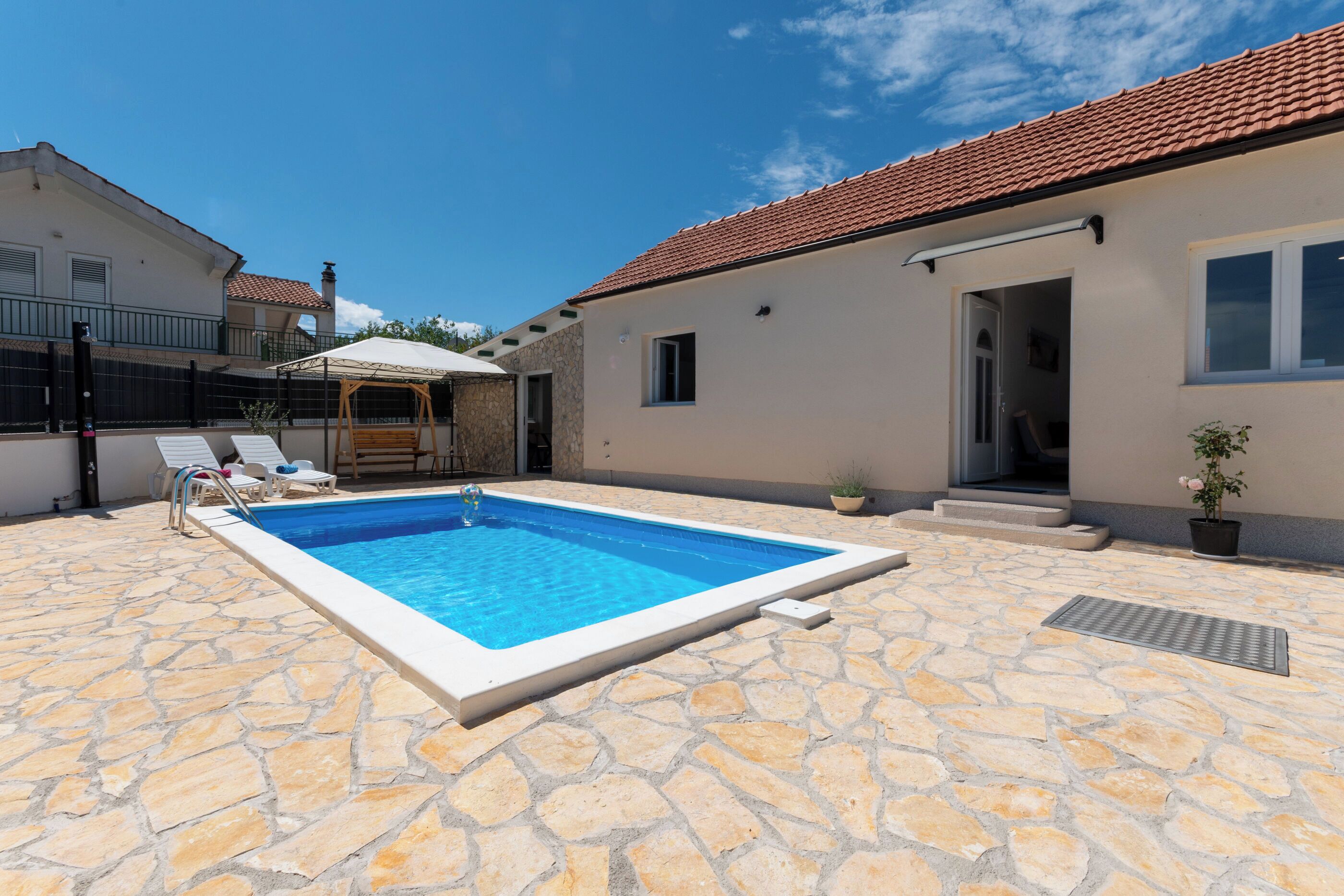 A swimming pool with stone flooring, a covered area with lounge chairs, and a building with a tiled roof.