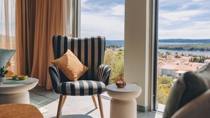 A room with a large window, a striped armchair with a cushion, a small table with a candle, and a view of a body of water and buildings. - Apartments Menigo Manzoni (Punat)
