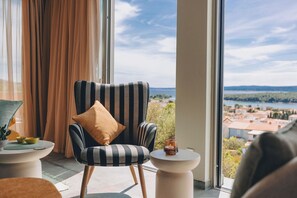 Ein Zimmer mit einem großen Fenster, einem gestreiften Sessel mit Kissen, einem kleinen Tisch mit Kerze und Blick auf ein Gewässer und Gebäude.