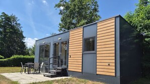 A modern single-story building with a combination of glass and wooden exterior, a patio with outdoor seating, and a clear sky. - Cube chalet voor 4 personen | De Otterberg (Wijster)