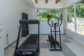 A home gym with a treadmill, elliptical, and weight machine, large windows with a view of greenery, and a stone wall. - Apartment Mediterranean (Opatija)