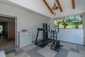 Ein Fitnessraum mit Laufband, Ellipsentrainer und Gewichten, einem weißen Schrank und einem Fenster mit Blick auf Grünflächen.