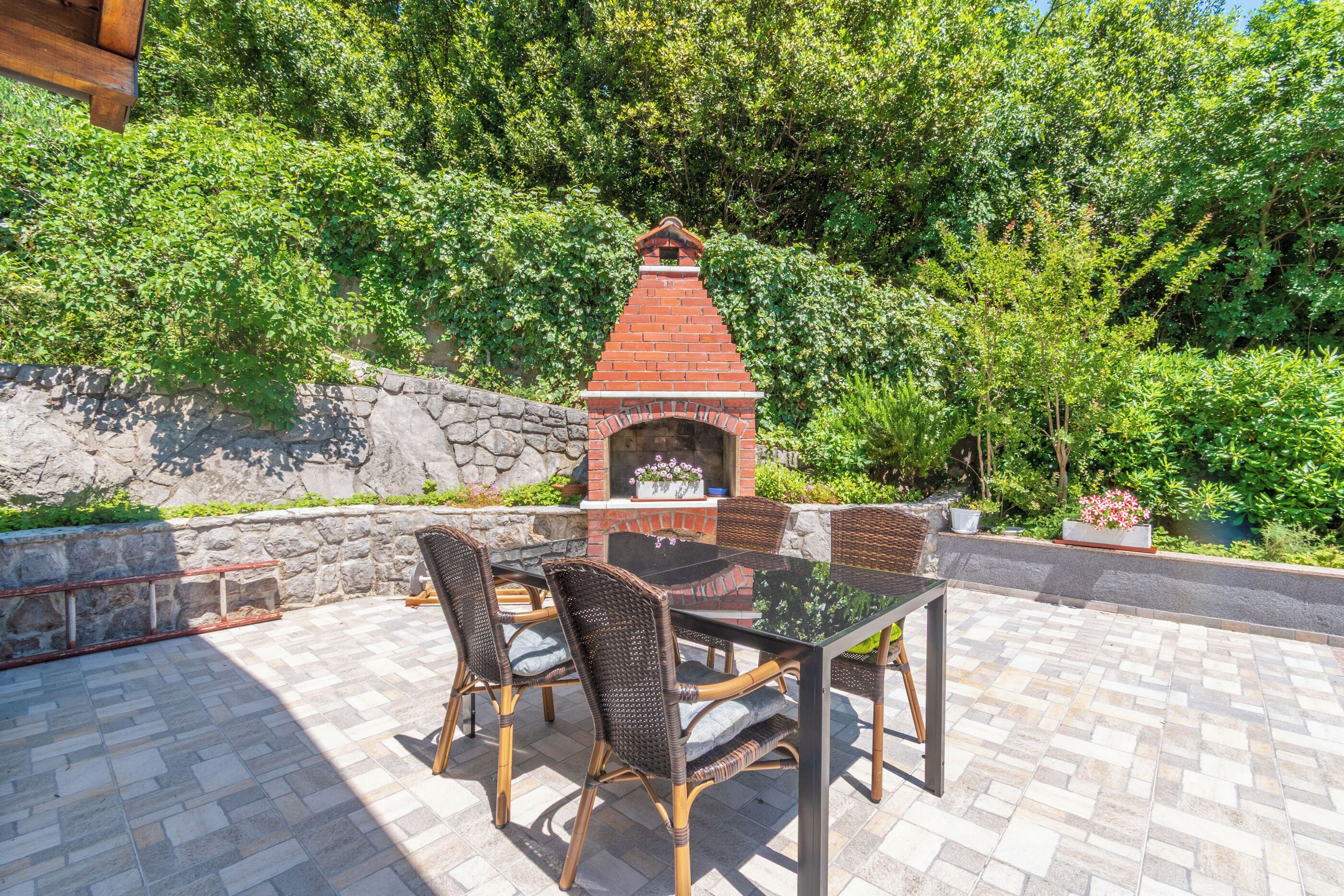 Eine Terrassenausstattung mit gemauertem Grill, Korbstühlen und einem Glastisch.
