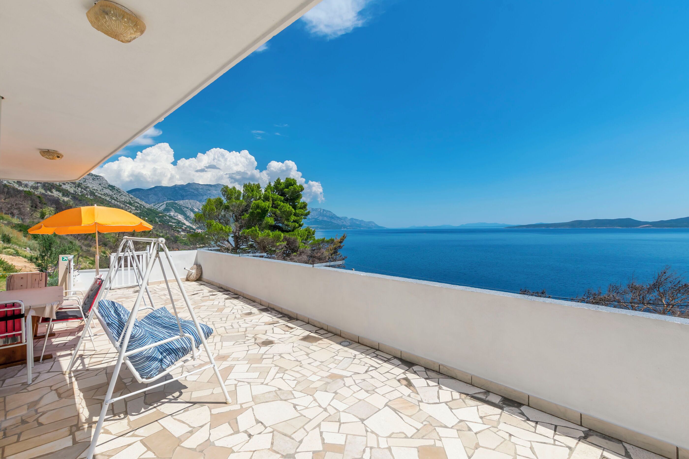 A terrace with a swing chair, a table, and an umbrella, overlooking a sea and mountains.