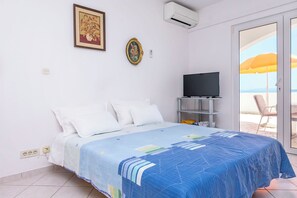 Ein Schlafzimmer mit Bett, Fernseher auf einem Schrank, einem Gemälde und Meerblick durch eine Glastür.