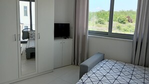 Ein Schlafzimmer mit einem Bett, einem Fernseher auf einer Kommode, einem Kleiderschrank und einem Fenster mit Blick auf Grünflächen.
