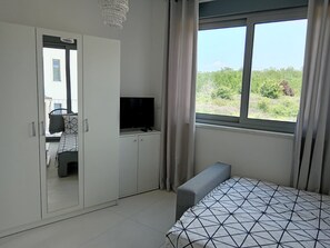 Ein Schlafzimmer mit einem Bett, einem Fernseher auf einer Kommode, einem Kleiderschrank und einem Fenster mit Blick auf Grünflächen.