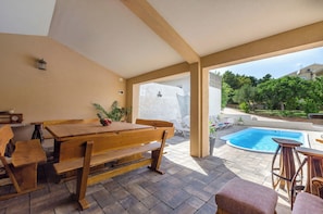 Ein Holztisch und Bänke in einem überdachten Außenbereich, im Hintergrund ein Pool.