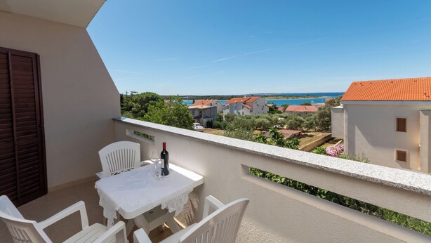 A balcony with a table and chairs, overlooking a coastal area with buildings and greenery. - Apartments Jadranka sea View (Novalja)