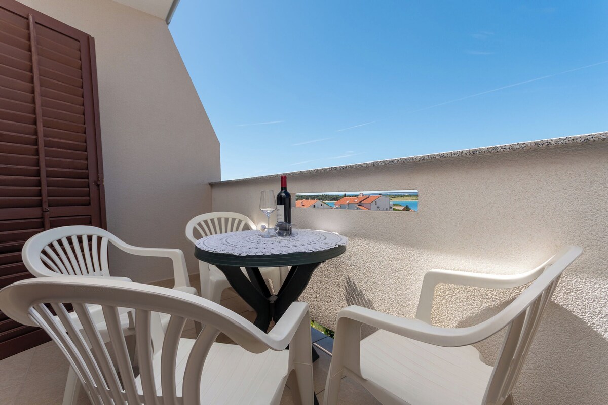 A small outdoor seating area with a round table and four white chairs, a bottle of wine, and a painting on the wall.
