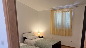 Um quarto com duas camas de solteiro, uma mesinha de cabeceira com abajur, uma janela com cortinas amarelas e piso de madeira.