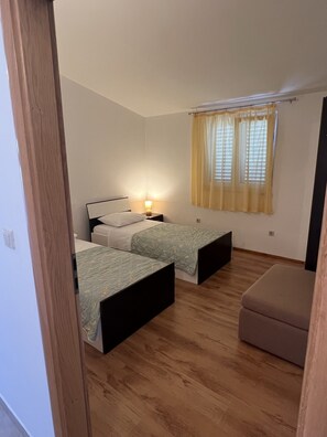 A bedroom with two single beds, a bedside table with a lamp, a window with yellow curtains, and a wooden floor. - Apartments Stipan (Marina)