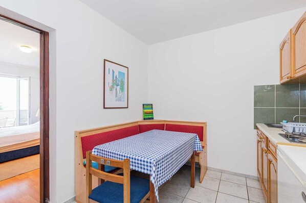 Una habitación compacta con una cama pequeña, una mesa con mantel a cuadros, un cuadro en la pared y una kitchenette con salpicadero verde.