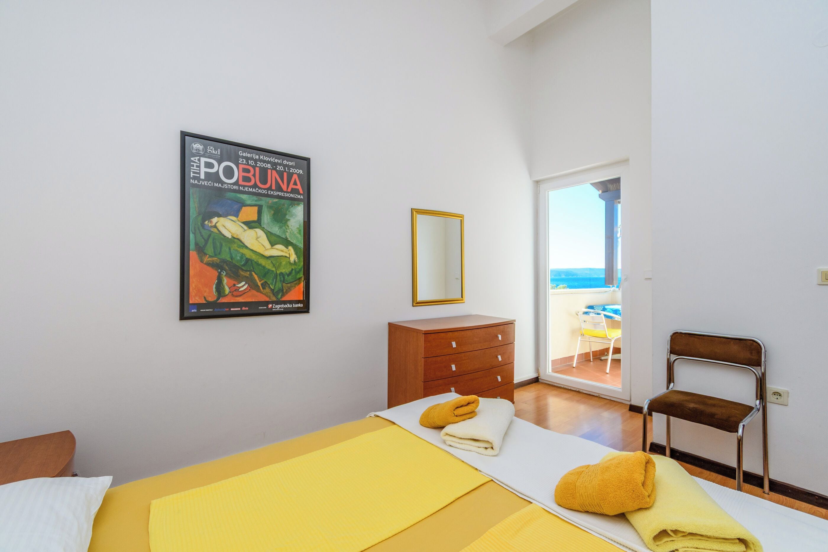 A hotel room with a bed, a wooden dresser, a chair, and a poster on the wall.