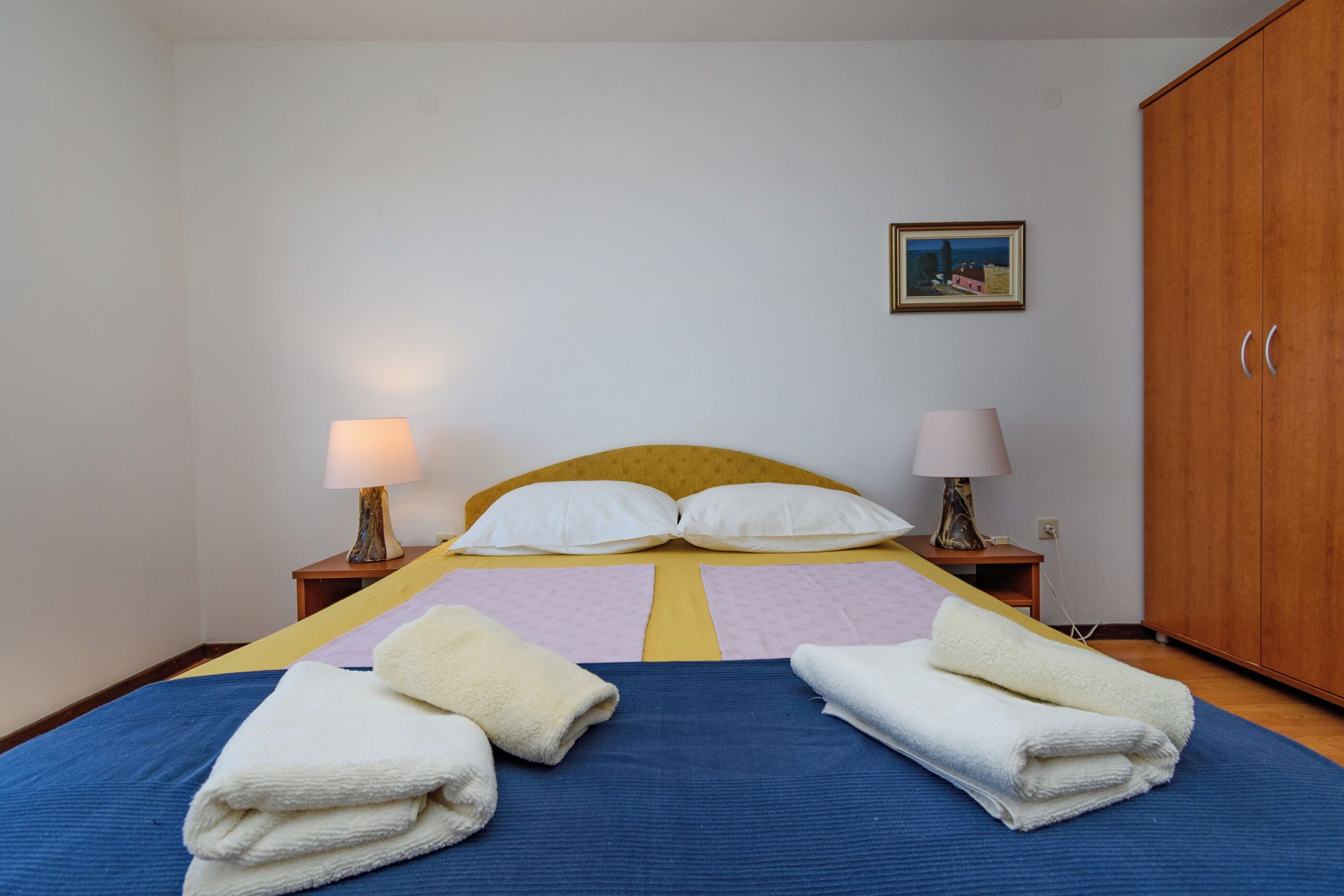 A bedroom with a bed, two bedside tables with lamps, a wardrobe, and a painting on the wall.
