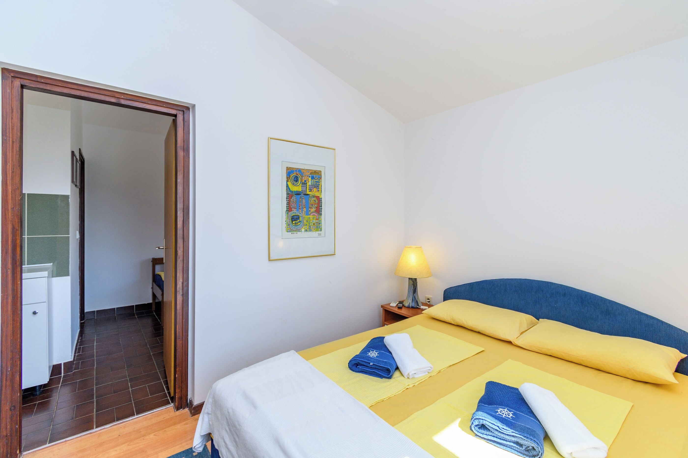 A hotel room with a bed, a bedside table with a lamp, a framed picture on the wall, and a bathroom visible through an open door.
