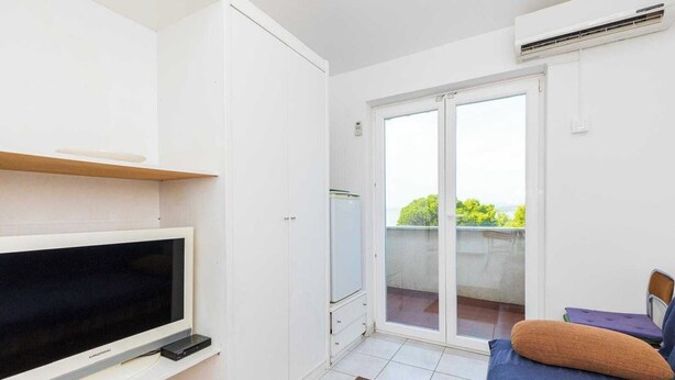Ein modernes Zimmer mit Flachbildfernseher, einer Schiebeglastür zu einem Balkon und einer kleinen Sitzecke mit Kissen.