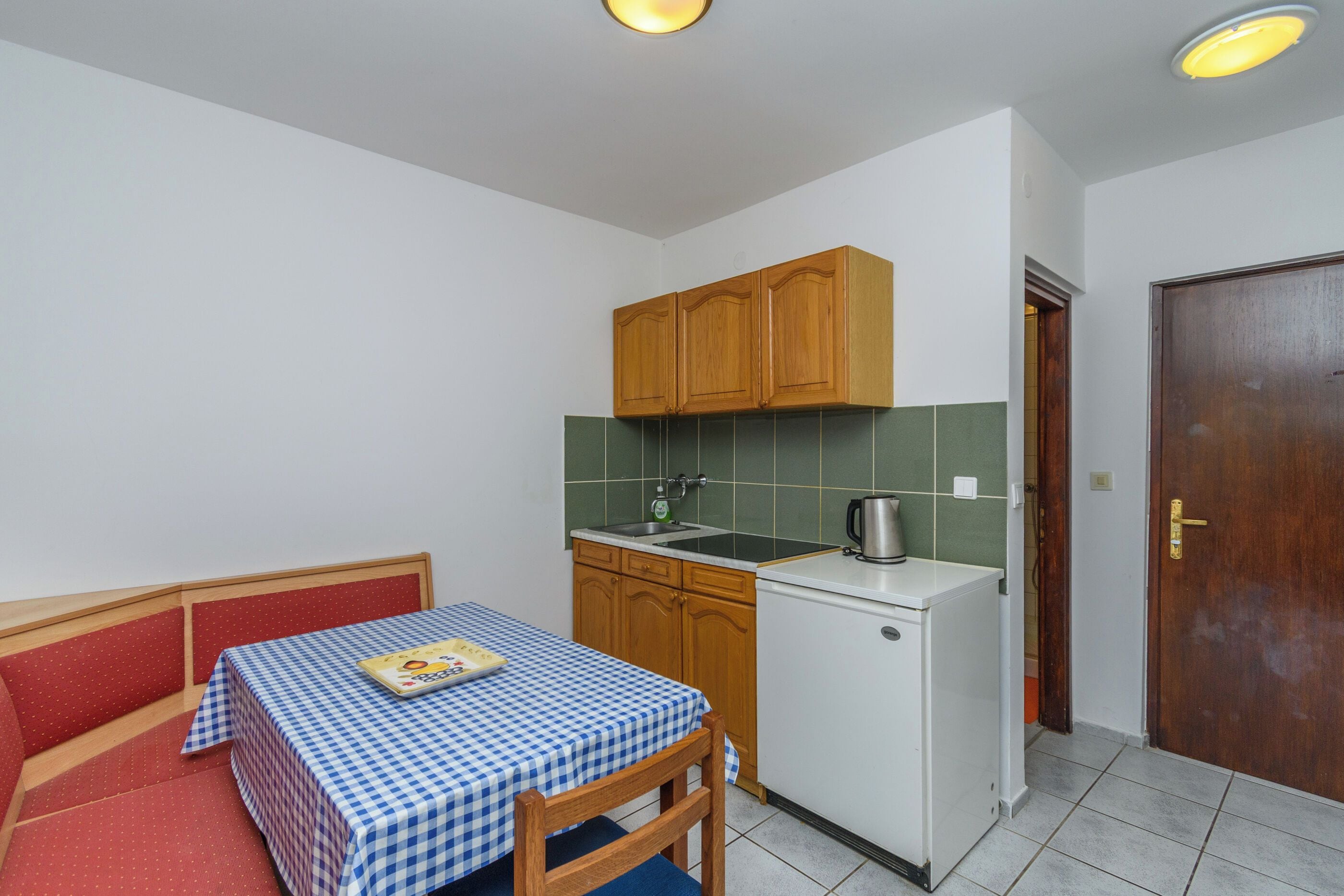 A compact kitchen with wooden cabinets, a small sink, and a refrigerator. There is a dining table with a checkered tablecloth and a chair.