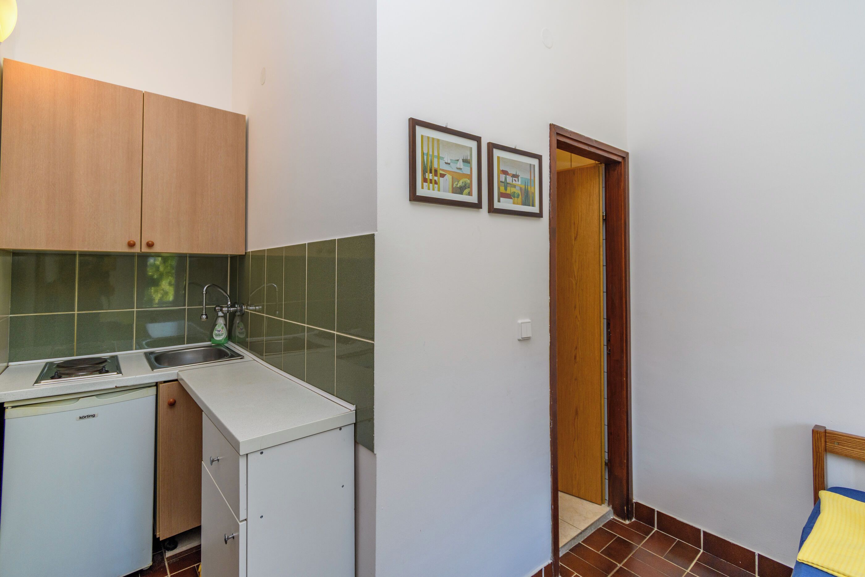 A compact kitchen with a sink, a small refrigerator, and wooden cabinets. There are two framed pictures on the wall and a door leading to another room.