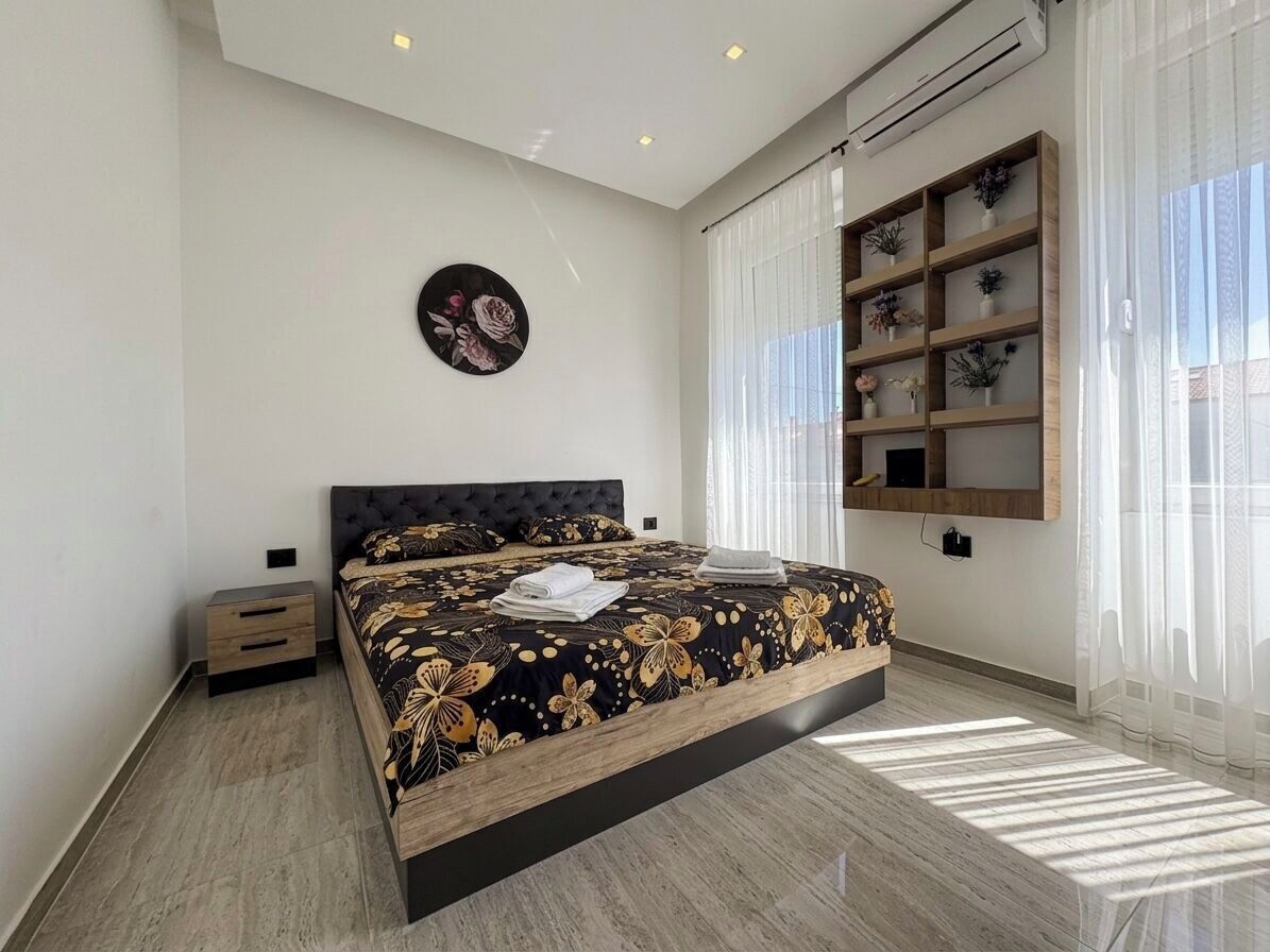 A modern bedroom with a large bed, a bedside table, a wall-mounted shelf with plants, and a window with sheer curtains.
