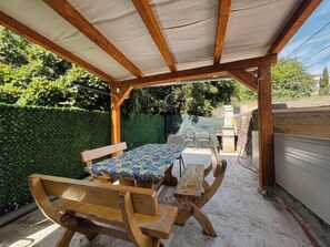 An outdoor patio with a wooden table and benches, a covered area, and a green hedge in the background. - House Holiday House Art (Crikvenica)