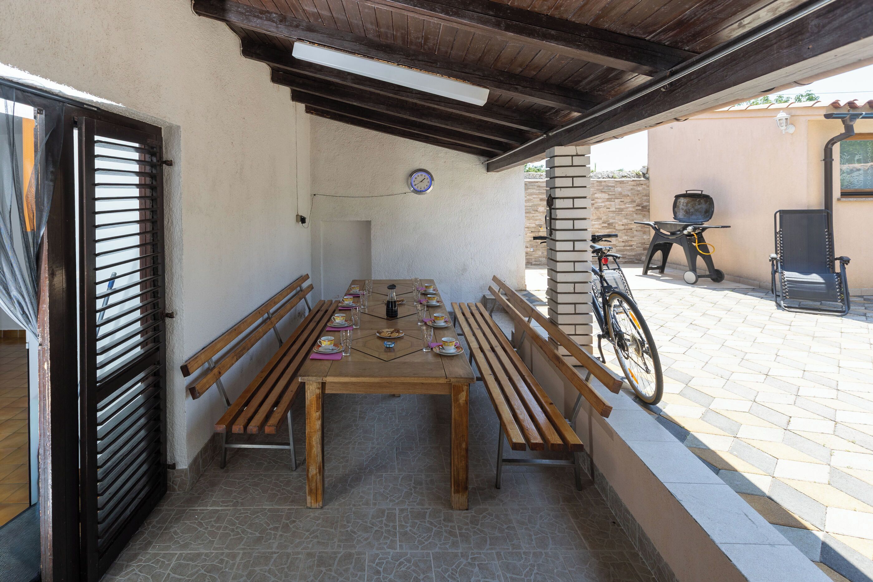 Eine Terrasse im Freien mit Holztisch, Bänken, einem Fahrrad und einem Grill.