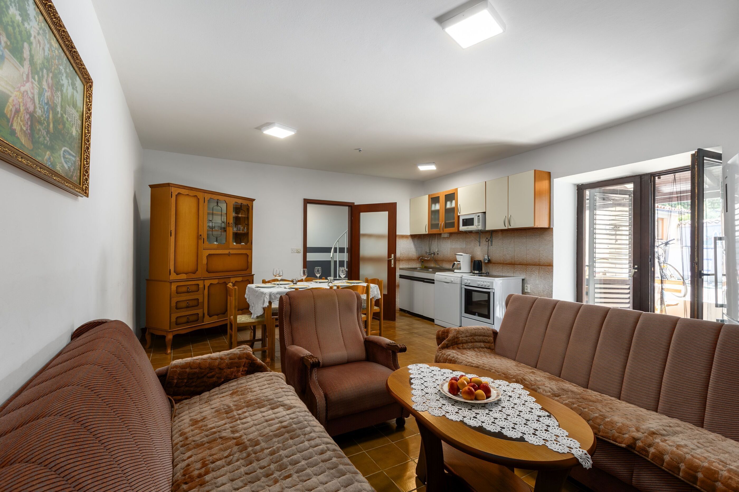 A living room with a sofa, armchair, dining table, and kitchen area with cabinets and appliances.