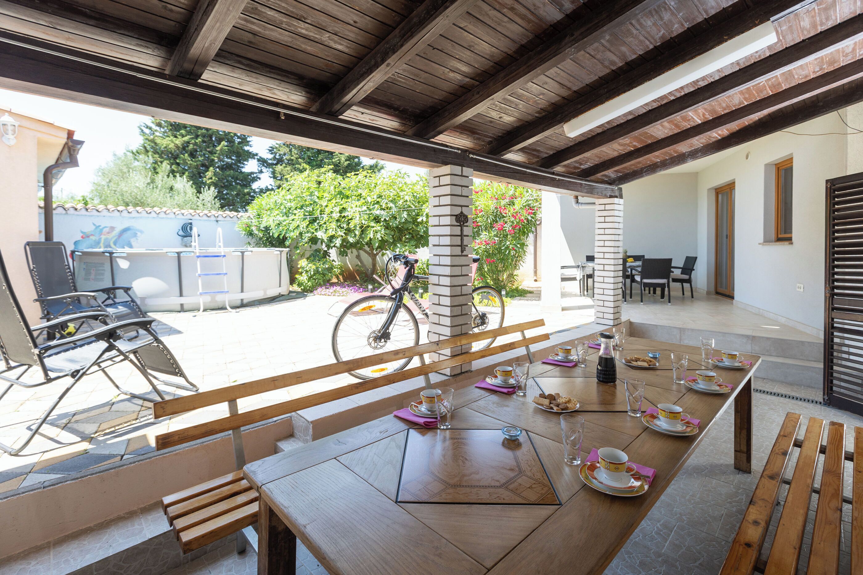 An outdoor patio with a table set for dining, a bicycle, and a view of the street and greenery.