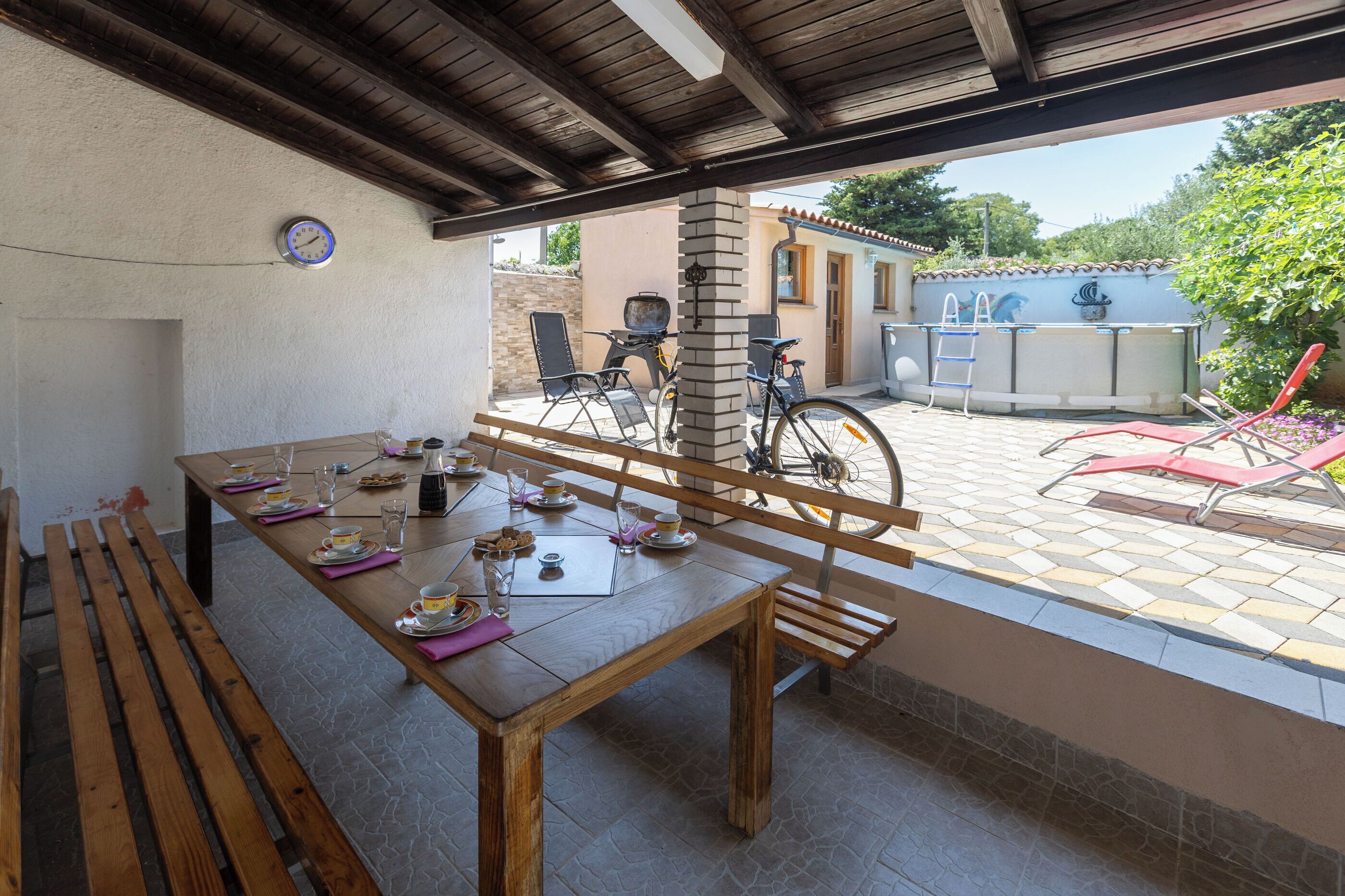 A covered outdoor dining area with a wooden table set for a meal, a bicycle, and a view of a building with a tiled roof.