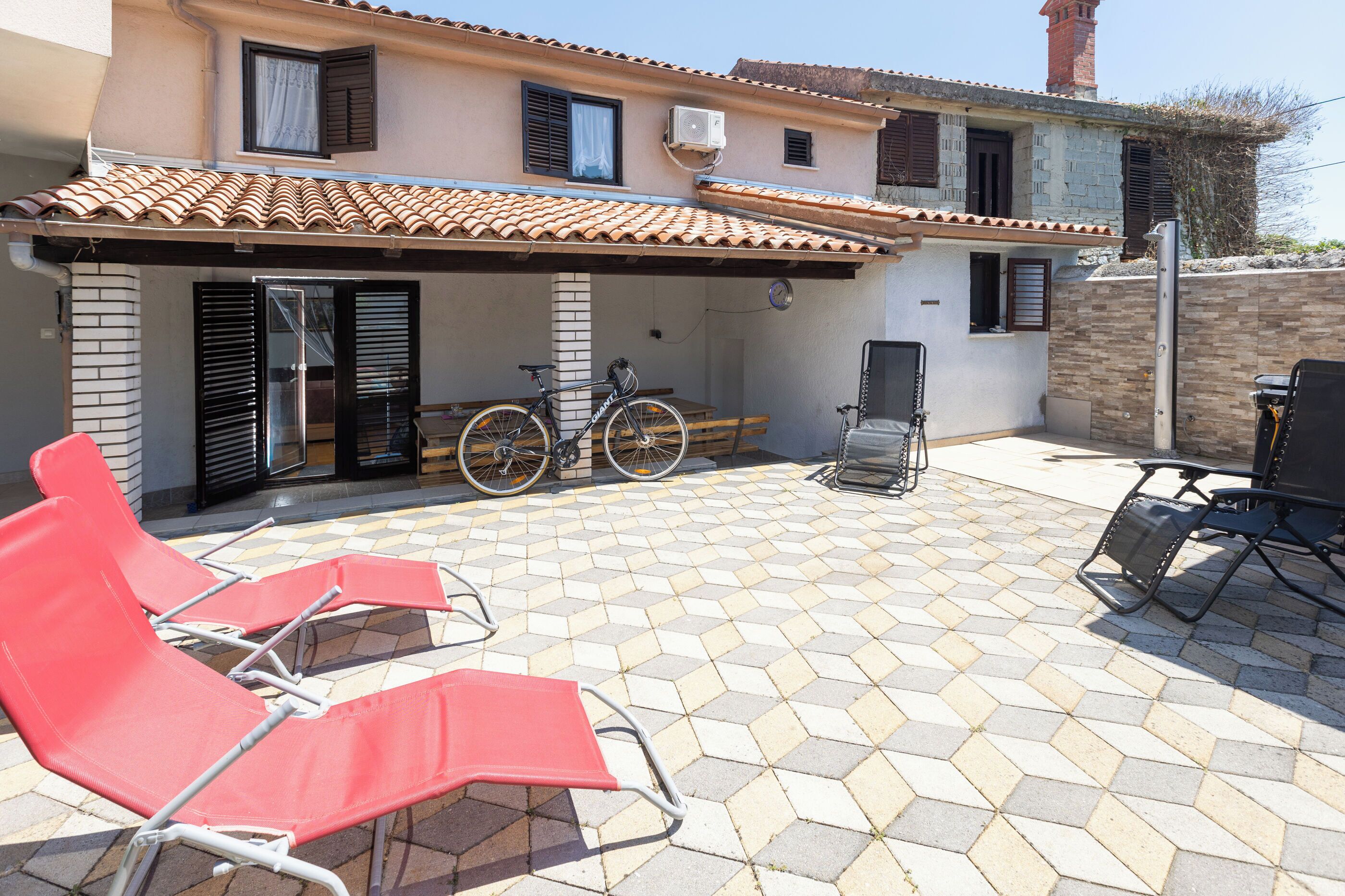 A covered outdoor area with a tiled floor, a bicycle, and red lounge chairs.